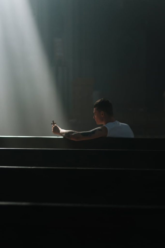 A contemplative moment in a church, as a man prays while holding a crucifix under a dramatic sunbeam.